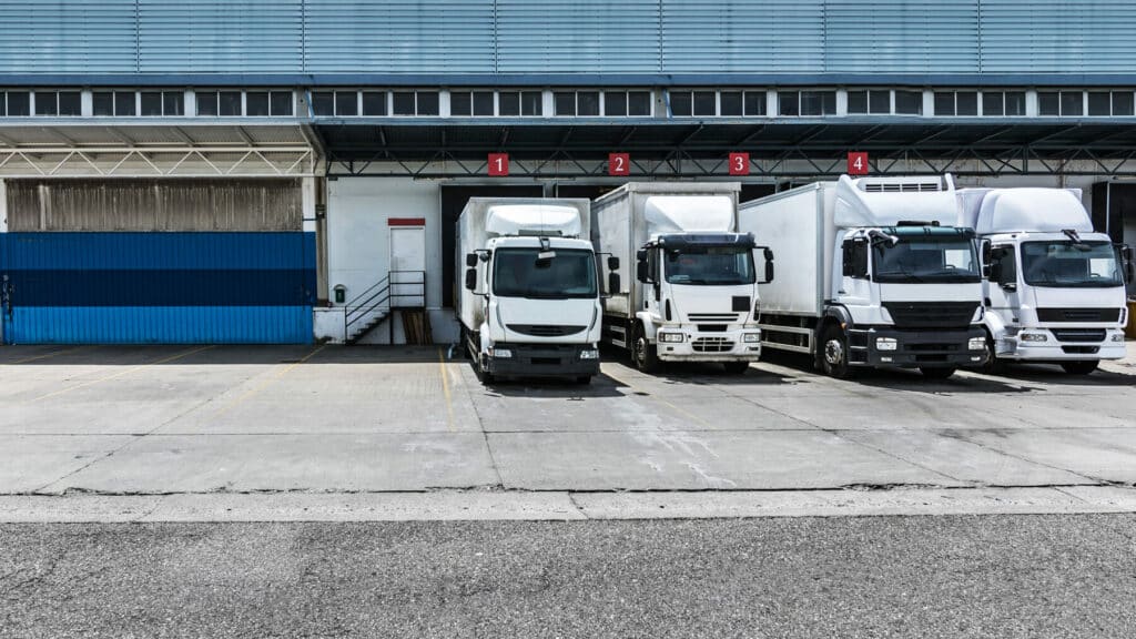 Vier schwere Lieferfahrzeuge stehen an nummerierten Verladerampen vor einem Logistikzentrum – sinnbildlich für die Neuwagenverkäufe, die unter die EU-CO2-Normen für schwere Nutzfahrzeuge (HDV) fallen