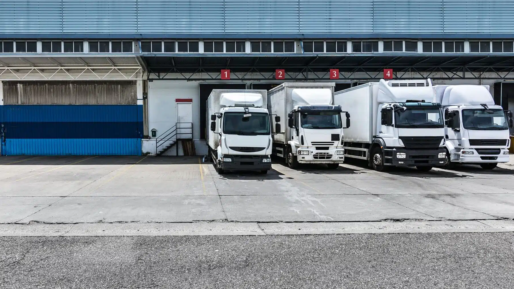 Four heavy-duty delivery trucks parked at numbered loading bays outside a distribution warehouse, representing the new-truck sales covered by the EU's HDV CO2 Standards Regulation