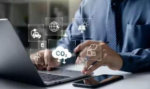 A business professional working on a laptop with digital icons overlaid showing CO2 emissions, a factory, renewable energy, and an electric vehicle, representing the digital tracking behind corporate emissions reporting