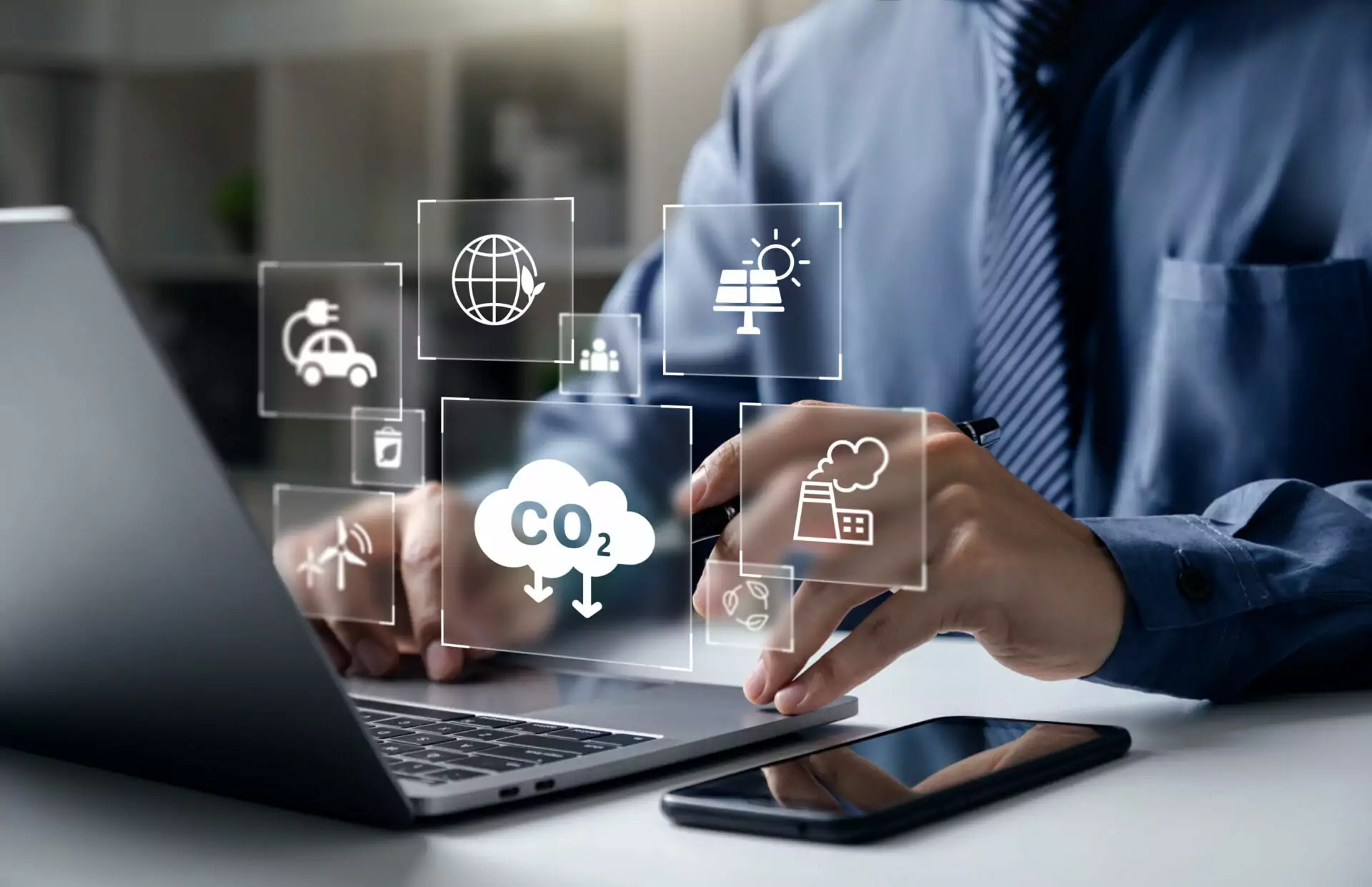 A business professional working on a laptop with digital icons overlaid showing CO2 emissions, a factory, renewable energy, and an electric vehicle, representing the digital tracking behind corporate emissions reporting