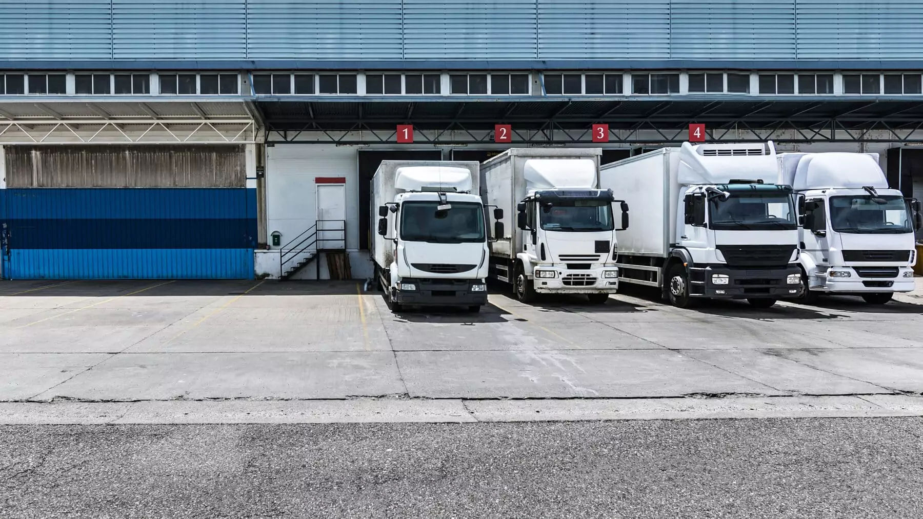 Four heavy-duty delivery trucks parked at numbered loading bays outside a distribution warehouse, representing the new-truck sales covered by the EU's HDV CO2 Standards Regulation